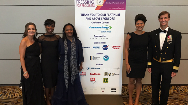 Four women in evening gowns and a man in a U.S. Navy officer dinner dress uniform stand other either side of a sign listing the platinum sponsors of the 2019 Women of Color in STEM conference. Sponsors include NOAA.