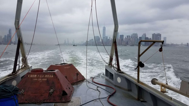 Two large red-colored sensors sit on the deck of a ship.The New York City skyline can be seen in the background. NOAA oceanographers deploy sensors like these in the water to measure the tidal currents for a period of time and then return to collect them. These two were recovered from the Hudson River, just off the coast of lower Manhattan.