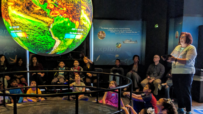An adult presents ocean current data on a Science on a Sphere to a group of children.