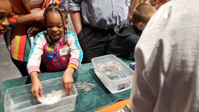 A child looks excitedly at their aluminum foil boat in a tub of water.