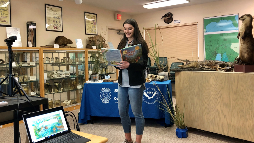 A teacher reads a children's book, The Secret Bay, in front of a web camera that is broadcast to elementary students in offsite classrooms. The educator is in a visitor center with plant and animal specimens and a map of the Gulf of Mexico.
