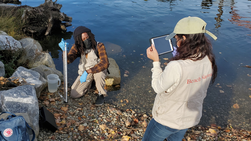 A naturalist is on their knee along a river edge with a turbidity tube, wearing a mask.  Another naturalist is using a tablet to record the activity. The river bank is made of cobblestones.