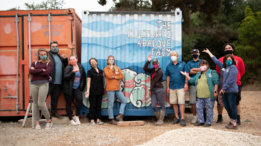 A group of people stand in front of a painted shipping crate. The crate says "The Cultured Abalone Farm."