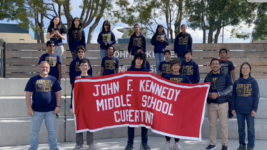 A group of students stand outside in school t-shirts while holding a sign that reads "John F. Kennedy Middle School Cupertino."