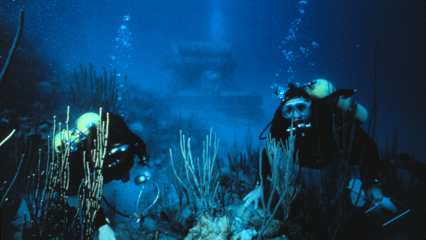 Two scientists in SCUBA gear float above a coral reef as they study it. The NOAA Hydrolab sits in the background