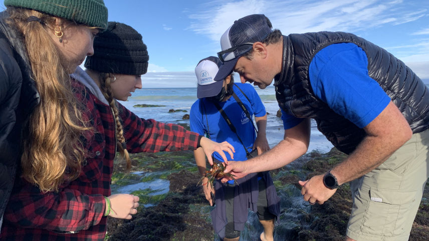 A male teacher holds a small kelp crab in his hand while several high school students look on in awe.