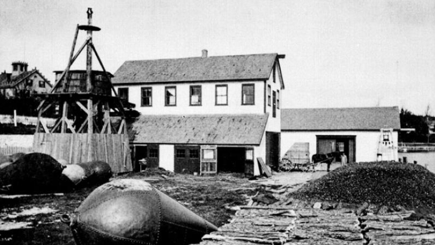 A black and white photo of the original Woods Hole, MA lab building with renovations, 1875. A crate housing a piano is visible on the horse-drawn carriage at right.
