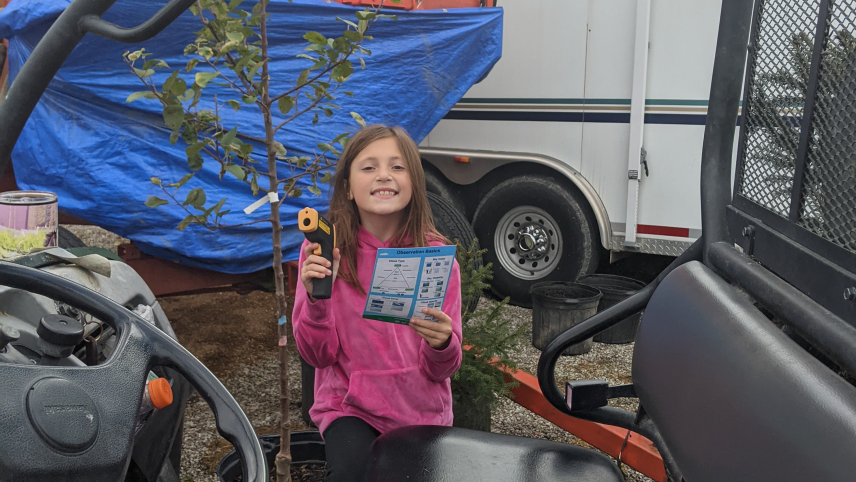 A an elementary student grins while holding up a temperature sensor and card with information about clouds. She is stepping up into a utility vehicle, and behind here are several small trees in pots. 