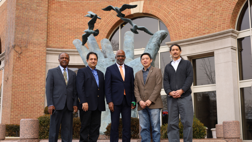 Five men in suits pose side by side in front of large statue of an open hand that appears to be releasing birds.