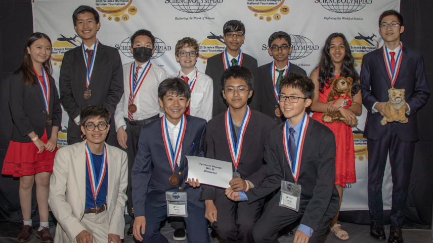Twelve students dressed in formal attire pose for a group photo in front of a backdrop that has the Science Olympiad National Tournament logo on it. The students in the front row are holding a sign that says Dynamic Planet Div B Winners!