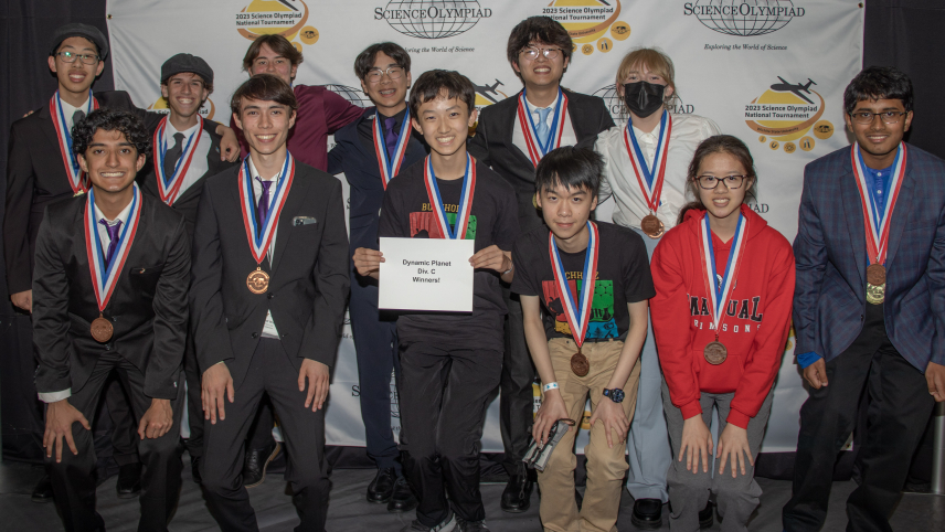 Twelve students dressed in formal attire pose for a group photo in front of a backdrop that has the Science Olympiad National Tournament logo on it. A student in the front row is holding a sign that says Dynamic Planet Div C Winners!