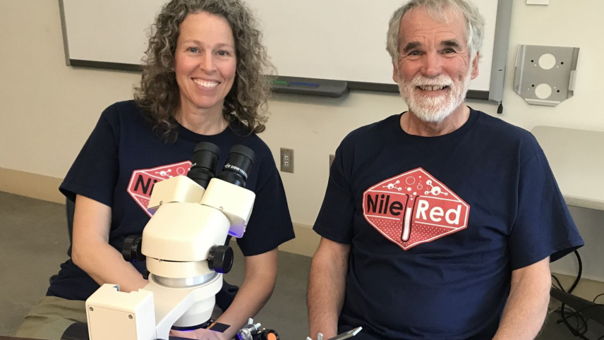 Clive Bagshaw and Amelia Labbe smile as they are sitting at a wooden table with a microscope that is also projecting a blacklight on the specimen. They are wearing matching blue t-shirts that have a red and white "Nile Red" logo.