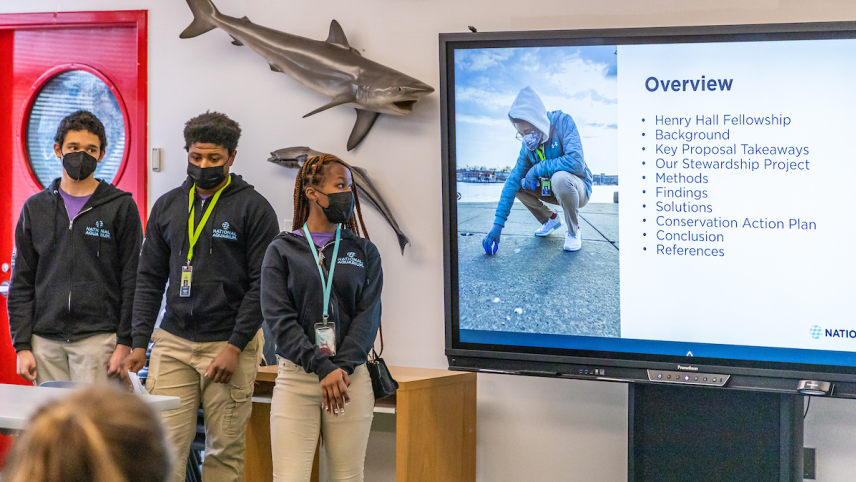 Three students wearing surgical masks and National Aquarium jackets stand in front of a screen presenting an overview of the project to an audience.
