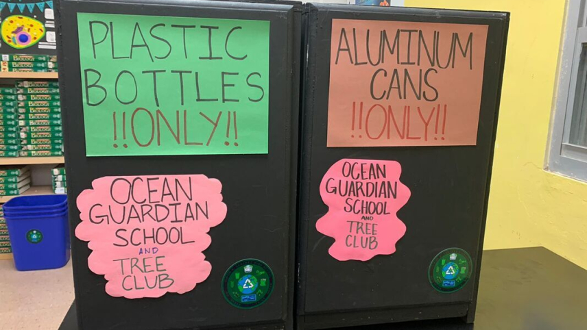 Two black waste bins are set up on a table for display. One has a large green sign with text reading "PLASTIC BOTTLES ONLY!!" and the other has a large orange sign with text reading "ALUMINUM CANS ONLY!!" Both also have a pink sign with text reading "Ocean Guardian School and Tree Club" and a blue recycling symbol. 