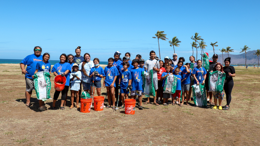 A group of about 30 people, mostly youth in matching t-shirts, on a beach. Many are wearing gloves or holding bags or buckets. Palm trees and a view of an ocean shore are visible behind them.