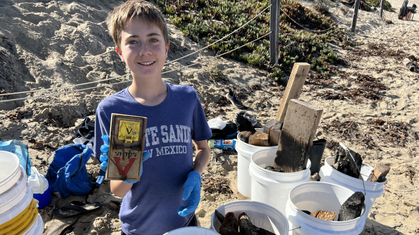 A young student wearing gloves kneels on a beach and holds up a discarded mousetrap. In front of them are several buckets filled with other marine debris.