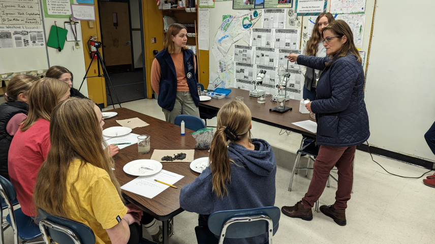 Three sanctuary educators stand at the front of a classroom with laboratory equipment, including microscopes, set up behind them as they teach about microplastics. 