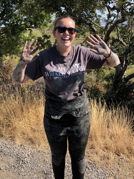 Carlee stands outside wearing a wet suit with the bib folded down, a short sleeve t-shirt, and is caked in mud. She grins and holds her muddy palms up for the camera.
