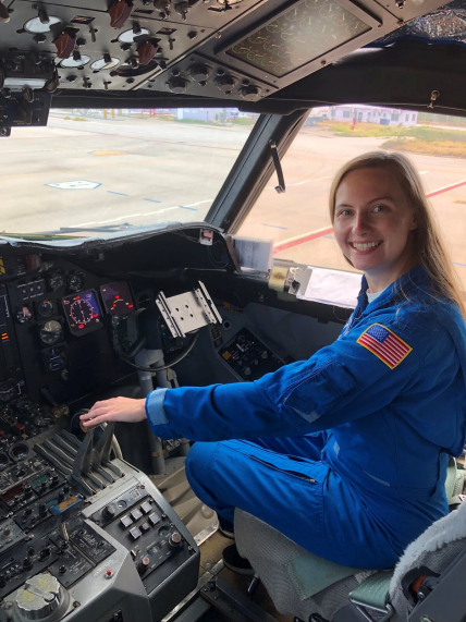  AOML Communications intern Holly Stahl smiling in an authentic blue NOAA flight suit while sitting in the cockpit of a P-3 Hurricane Hunter aircraft.