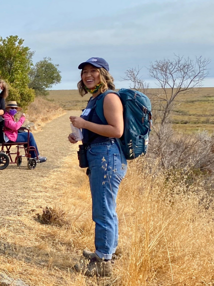 Bella Mayorga stands on a path overlooking a marsh while smiling. She is wearing a backpack and binoculars and appears to be speaking to a group of people, one of whom is visible in the background in a wheelchair.