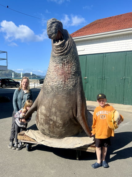 An adult and two children stand on either side of a statue of a large male northern elephant seal. A storage shed next to a parking lot is in the immediate background with a view of San Francisco Bay in the distant background.