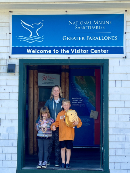 A child is holding a doll and another child is holding a stuffed animal in front of an adult. All three people are standing in front of a building with a sign that reads “National Marine Sanctuaries - Greater Farallones. Welcome to the Visitor Center.” 
