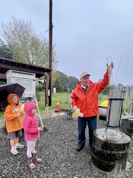 A child holding an umbrella next to another child in a jacket and rainboots stand to the left of an adult in a rain jacket and baseball cap holding a cylinder that measures the amount of water collected. The cylinder is suspended above a metal cylinder that sits on a larger metal cylinder.