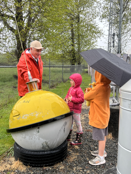 A child holding an umbrella next to another child in a jacket and rainboots stand to the left of an adult in a rain jacket and baseball cap holding a cylinder that measures the amount of water collected. The cylinder is suspended above a metal cylinder that sits on a larger metal cylinder. 