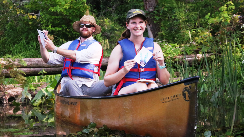 Two educators sit in a canoe, smiling and drawing in their notepads.