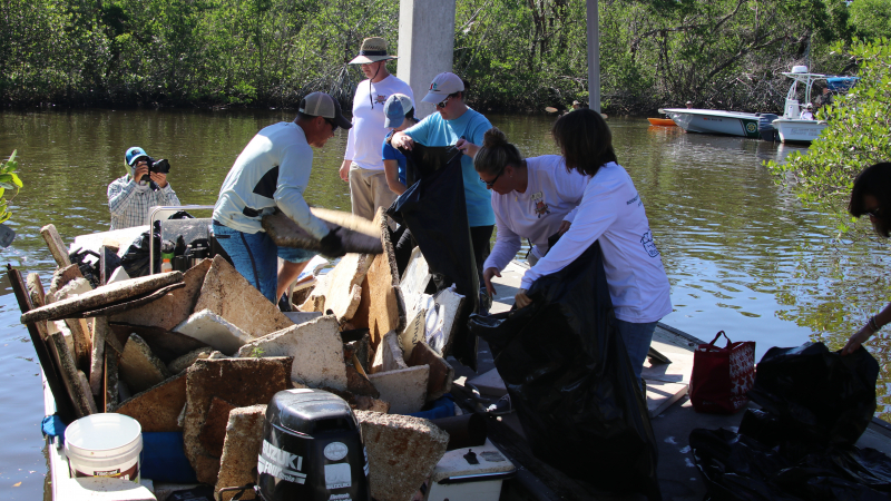 Volunteers unload a pile of large debris from a small motor boat onto a dock in a mangrove forest. 