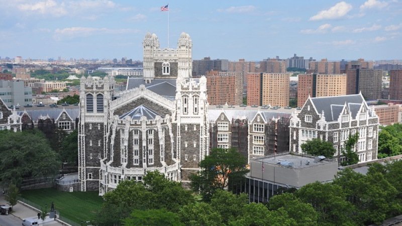 Several buildings in English Gothic style, built with dark stone and white trim. Trees surround the campus in the foreground and a large city sits behind the campus.