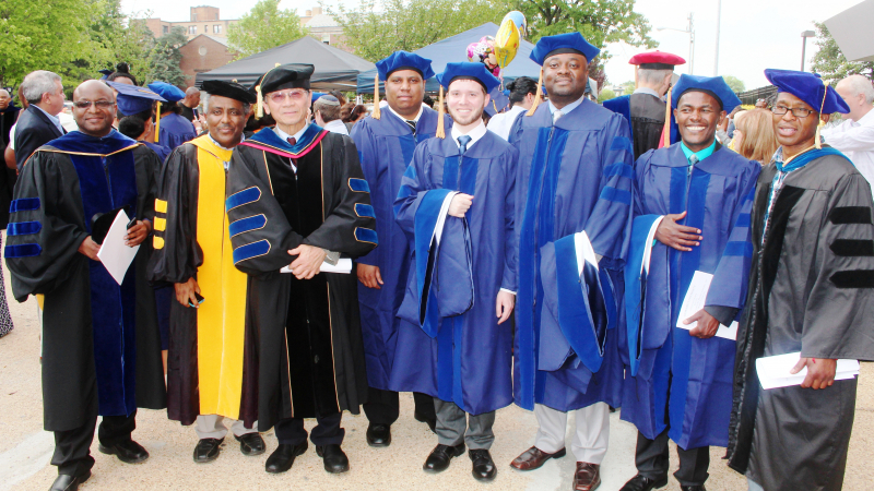 Students and professors stand together at graduation. Four are wearing Masters level robes, one is wearing newly graduated PhD robes and three are wearing professor robes.