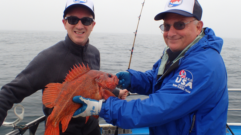 Two men stand together on a boat holding a large, red Vermilion rockfish.