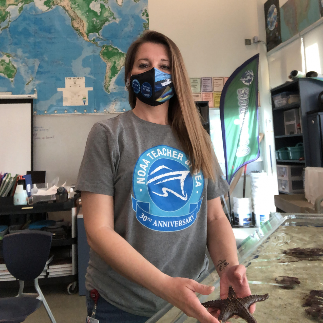 A teacher stands in a bright classroom wearing a cloth mask over her nose and mouth. She is holding a sea star from a touch tank.