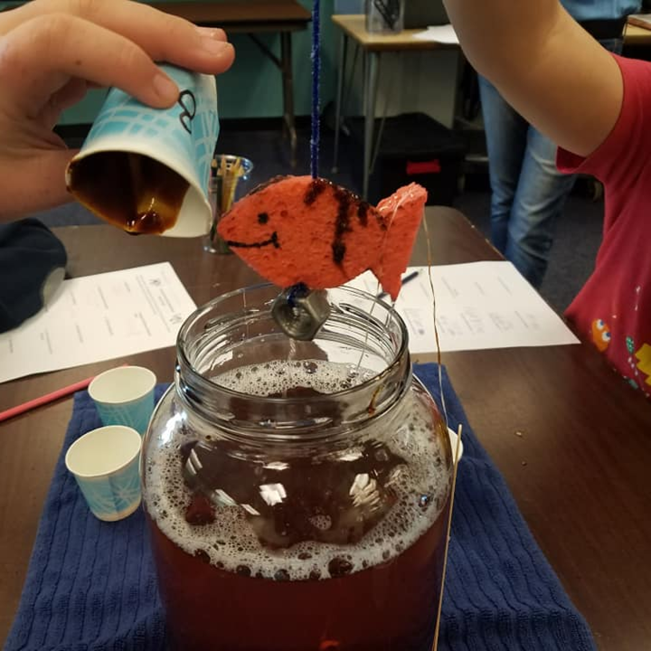 Student hands work with an activity that has a sponge shaped like a fish being dunked into a mason jar full of murky liquid.