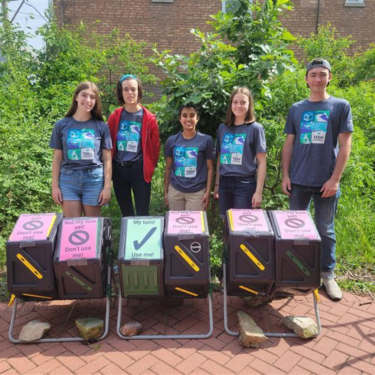 Five teens wearing matching aquarium t-shires stand behind a row of six plastic compost bins. Five of the bins have signs on top that say, "Not my turn yet! Don't use me!" while one says, "My turn! Use me!" They are outdoors, surrounded by lush greenery.