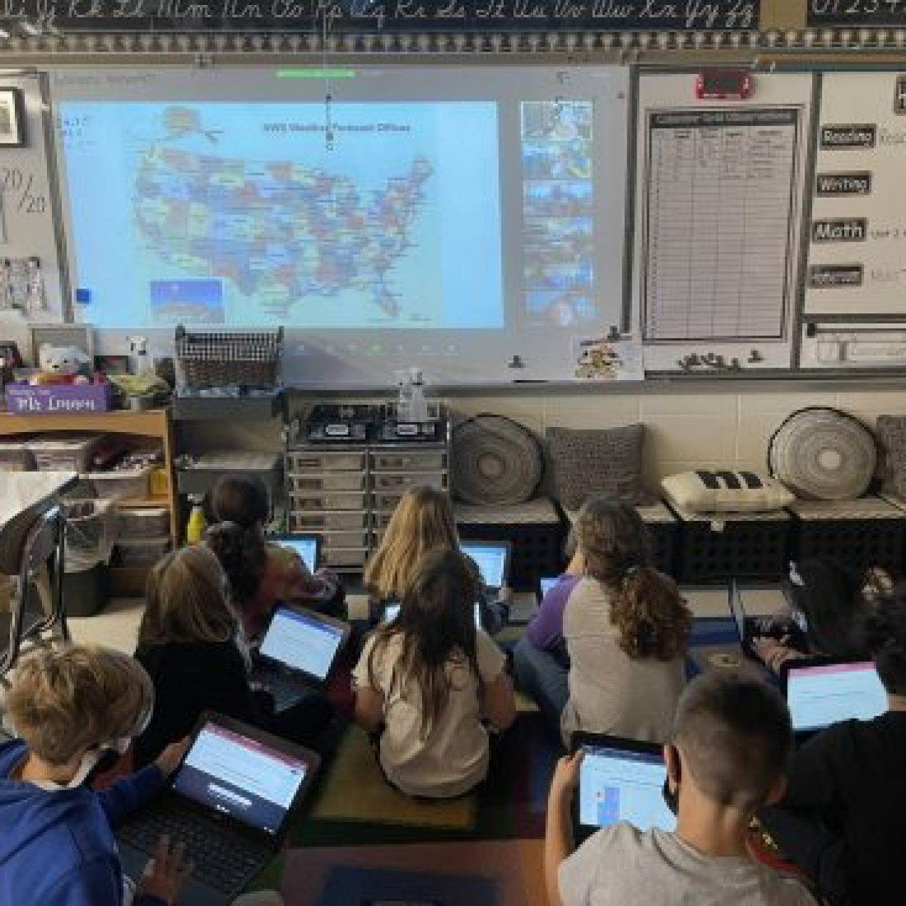 A classroom of students use iPads while following along with a virtual presentation in the classroom.