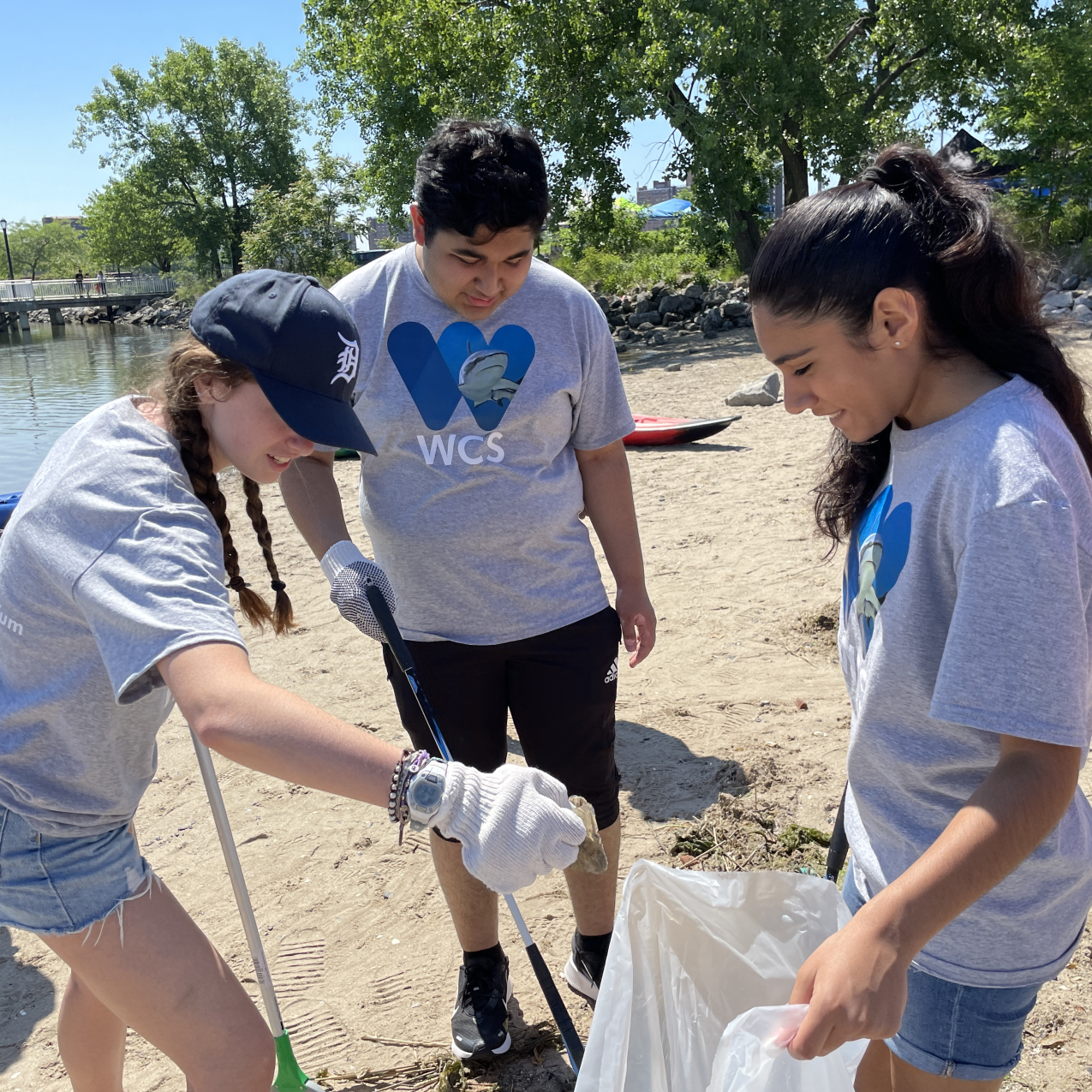 Three people are standing outside participating in a beach clean-up. The person on the right is holding a garbage bag open while the other two people are holding an object grabber. The person on the left is putting something into the garbage back with their gloved hand. In the background, there are trees and a body of water.