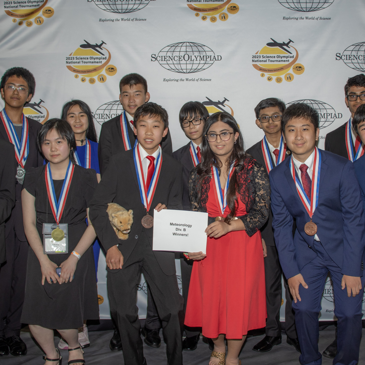 Twelve students dressed in formal attire pose for a group photo in front of a backdrop that has the Science Olympiad National Tournament logo on it. The students in the front row are holding a sign that says Meteorology Div B Winners!
