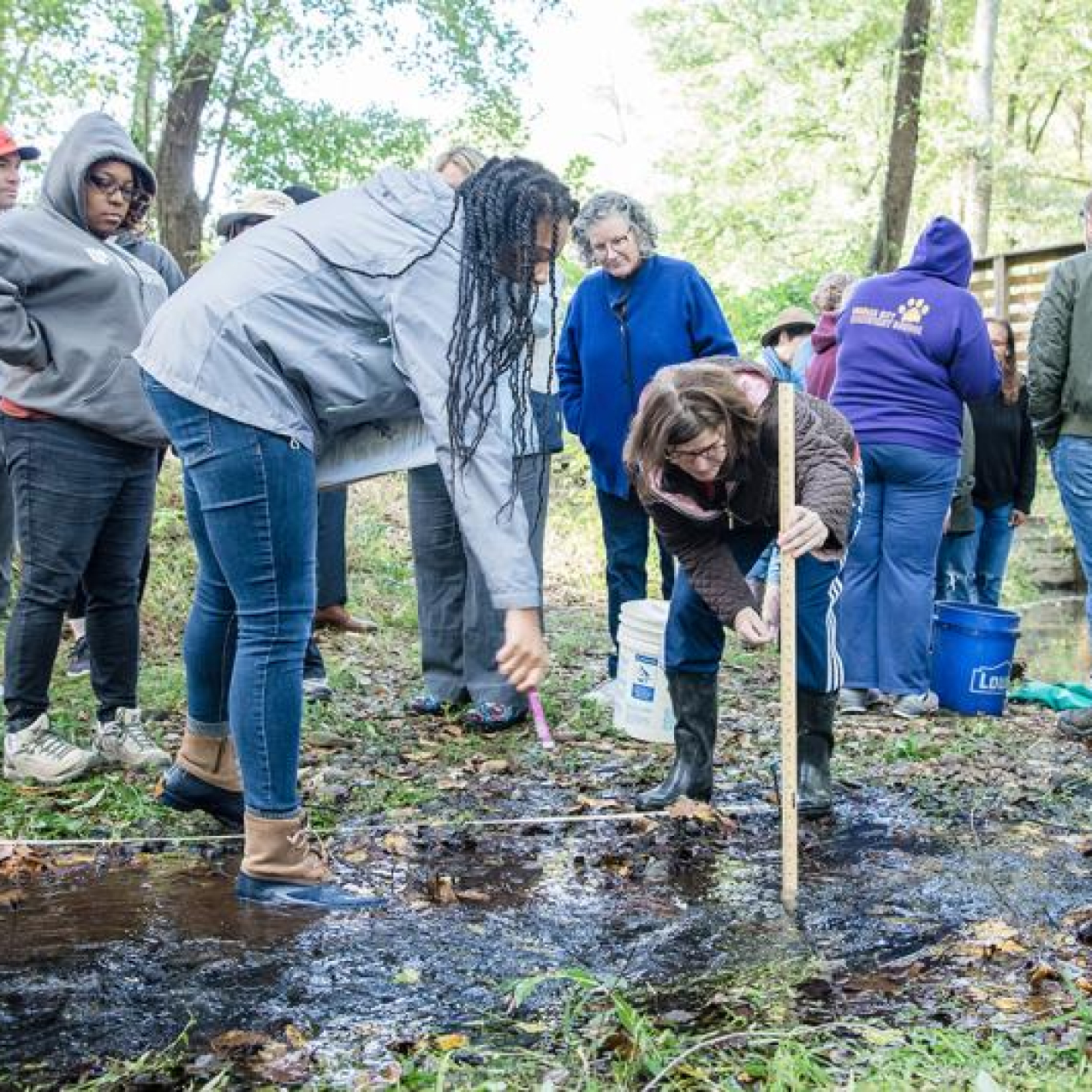 A group of educators stand together outside with buckets, meter sticks and other sampling supplies. The educators are participating in environmental education and look to be measuring a shallow puddle.