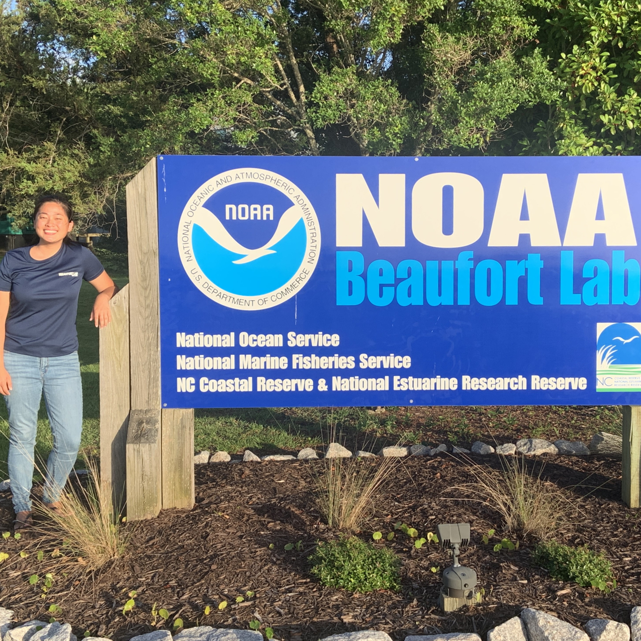 Michaela stands next to a large sign that says "NOAA Beaufort Lab" beside a NOAA logo. Smaller text reads "National Ocean Service, National Marine Fisheries Service, NC Coastal Reserve and National Estuarine Research Reserve