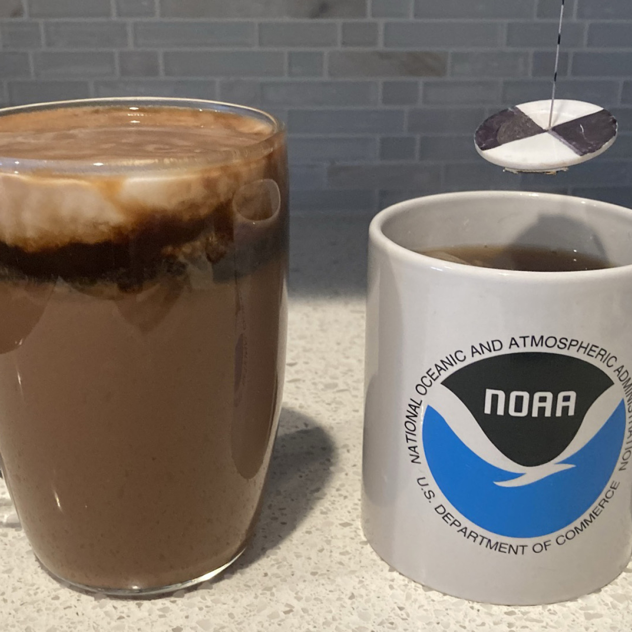 Learn about density by making a fancy layered hot cocoa or find out the "secc-tea" depth of your perfect up of tea with our oceanography in a mug activities. 
Two mugs are placed on a kitchen counter. One mug has the NOAA logo on it and a miniature secchi disk is suspended above it. (The secchi disk is made of a white disk with black on opposite quarters. It is hanging from a string that has markings every centimeter.) The other mug is made of clear glass, and it contains hot cocoa topped with a layer of