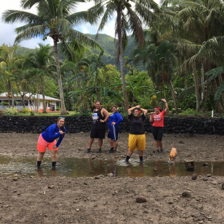 Five college students and a dog pose in and around a stream in American Samoa. Palm trees and mountains are in the background.