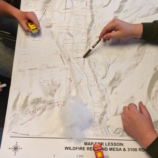 A topographic map of Redlands Mesa on a table with several students' hands visible. One student indicates an area with a marker. The wind direction has been drawn on the map with an arrow, and students appear to be using toy fire trucks and cotton batting that resembles smoke in a planning exercise. 