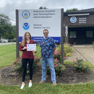Lillian and Ryan stand next to each other in front of a National Weather Service building and sign that reads "National Oceanic and Atmospheric Administration, National Weather Service, Louisville Weather Forecast Office." Lillian is holding a certificate of recognition.  