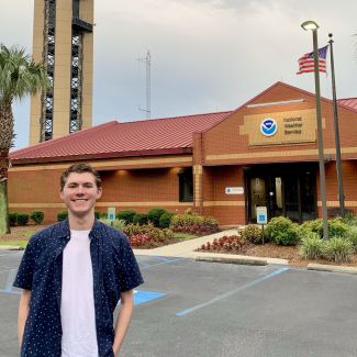 Connor poses in front of a building that has a NOAA logo and the words "National Weather Service" above the door. 