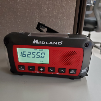 Small black NOAA weather radio made by Midland with red and black housing, an antenna, and six control buttons. 