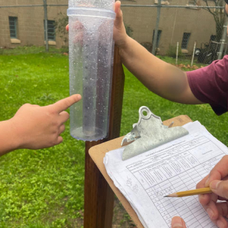 A rain gauge on a post in a grassy outdoor area surrounded by a chain-link fence. Just the arms and hands of students are visible as one student holds the gauge, one points to indicate the level of rainfall that has collected in the gauge, and a third records information on a data sheet. 