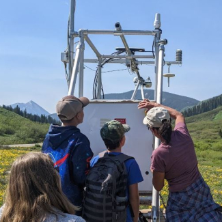 Three children and an adult examine a large metal structure mounted with electronic devices. They are standing in a wide alpine valley filled with wildflowers and surrounded by high mountains.