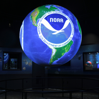 A Science on a Sphere in a dark room. The Sphere has a map of the world with a large NOAA logo on top.